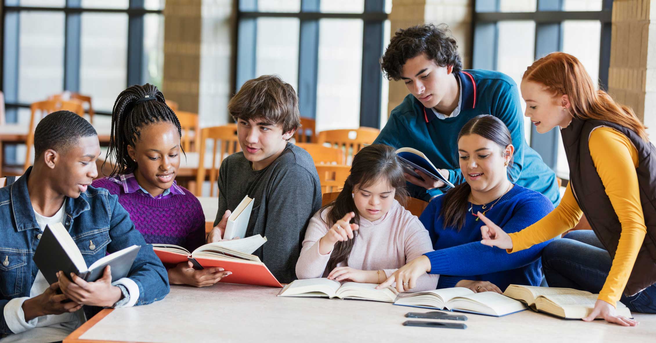 A group of students at a library table reading books and talking.